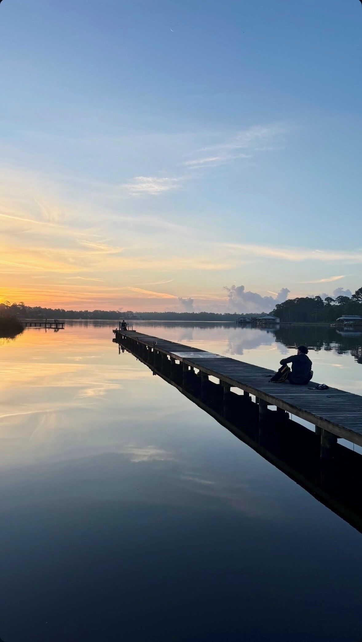 Um lago tranquilo com um píer de madeira que se estende sobre a água, uma pessoa sentada no píer e uma linha de costa distante com edifícios.