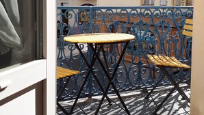 A balcony with a small table and two chairs, a wrought iron fence, and a building with a sign that reads "RESTAURANT" in the background.