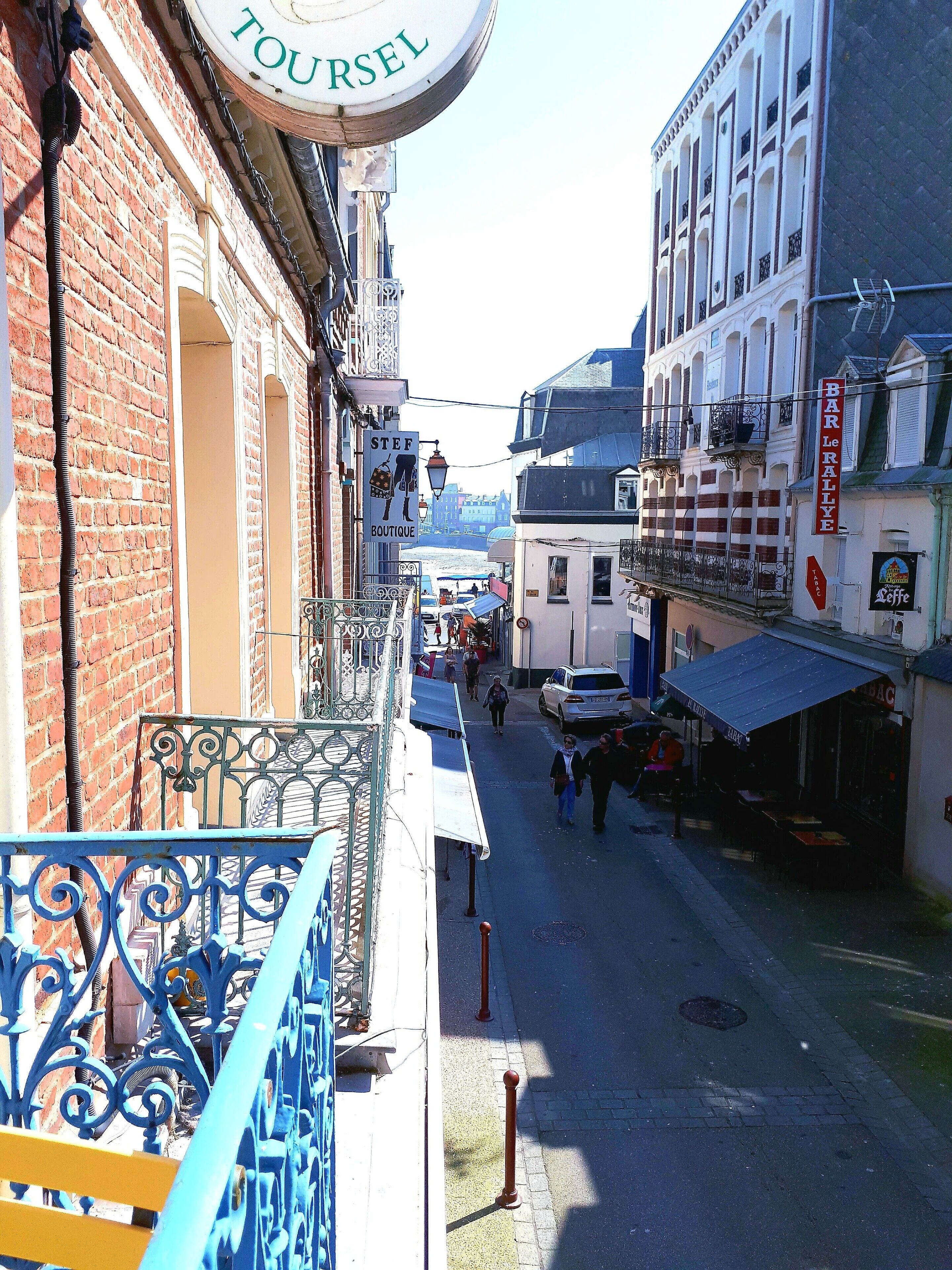 A narrow street with buildings on either side, a sign for 'TOURSEL' above a building, and a 'BAR Le RALLYE' sign on a building.