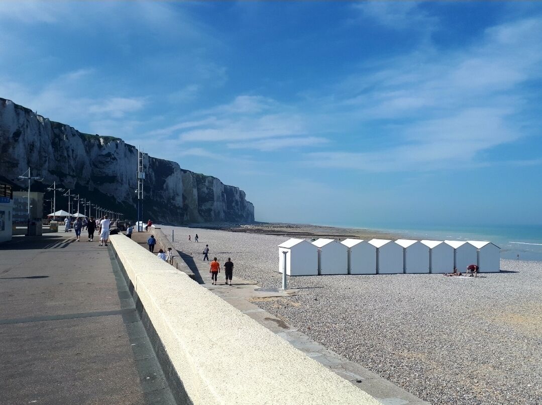 A coastal promenade with a rocky cliff, a pebble beach, and a row of white beach huts.