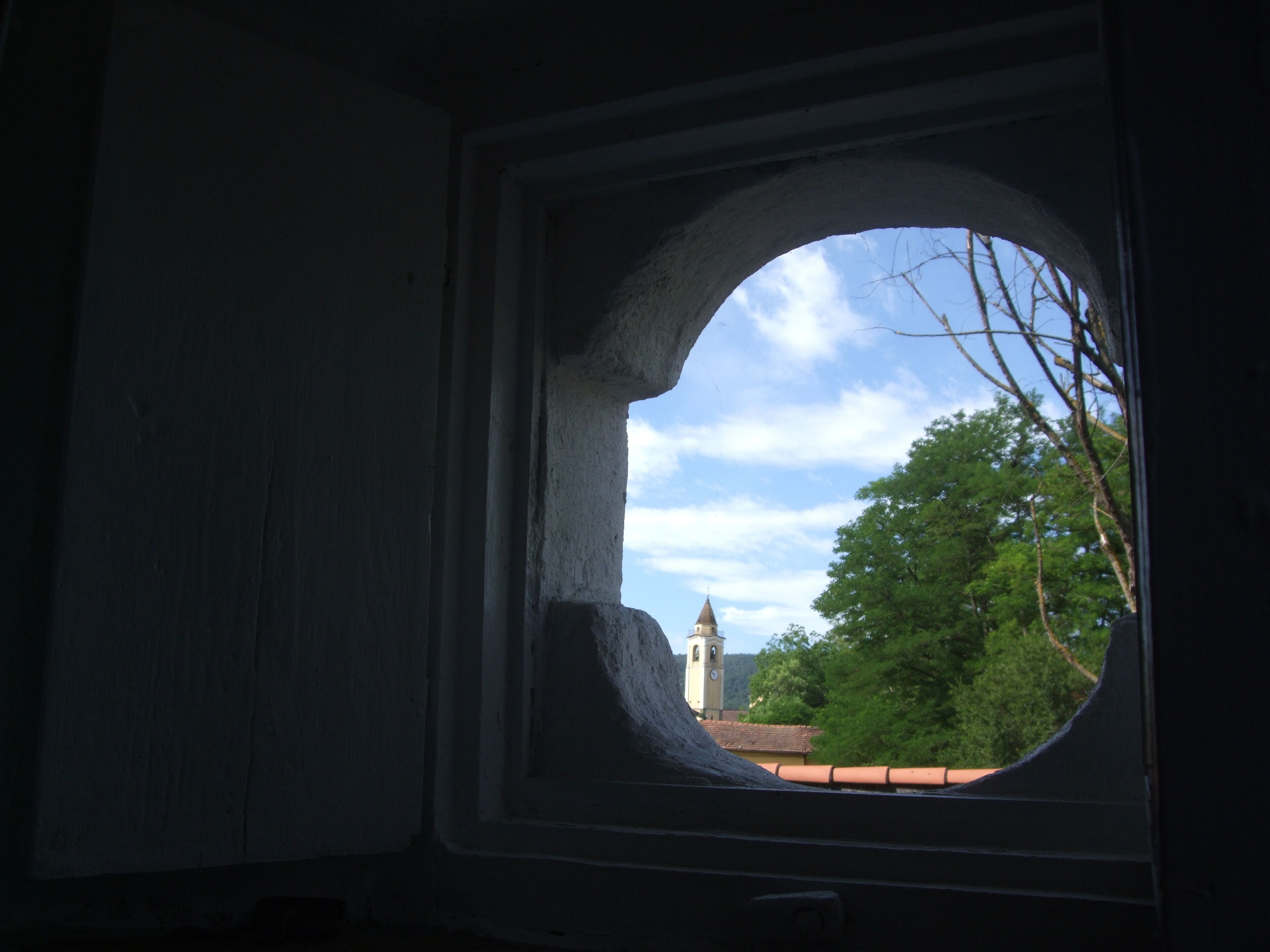 A view through a window with a stone arch, overlooking a building with a clock tower and trees.