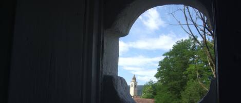 A view through a window with a stone arch, overlooking a building with a clock tower and trees.