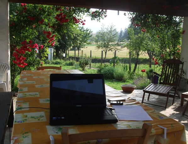 A laptop on a table with a yellow tablecloth, surrounded by chairs and a garden view.