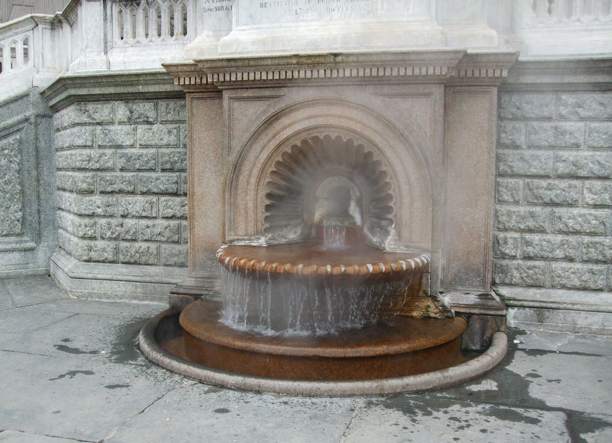 Une fontaine en pierre, dont l’eau jaillit dans un bassin circulaire, est adossée à un mur de pierre surmonté d’une arche décorative.