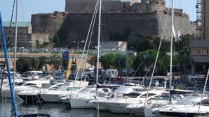 Un puerto deportivo con varias embarcaciones amarradas, una fortaleza histórica al fondo y un cielo despejado.