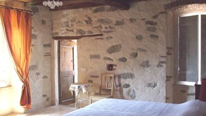 Una habitación rústica con pared de piedra, una cama de madera, una mesita con mantel y una ventana con cortinas.