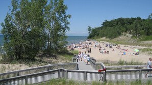 A beach with a wooden boardwalk leading to the shore, surrounded by trees and a clear sky.