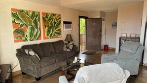 A living room with a sofa, armchair, and coffee table. There are two large paintings on the wall, a door with a number, and a bookshelf with books. - Stunning Ocean Views Steps from Sugar Beach (Kihei)