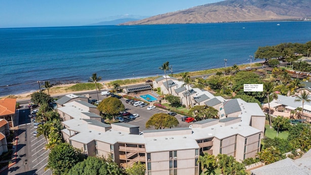 A coastal hotel complex with multiple buildings, a parking area, and a view of the ocean. - Stunning Ocean Views Steps from Sugar Beach (Kihei)