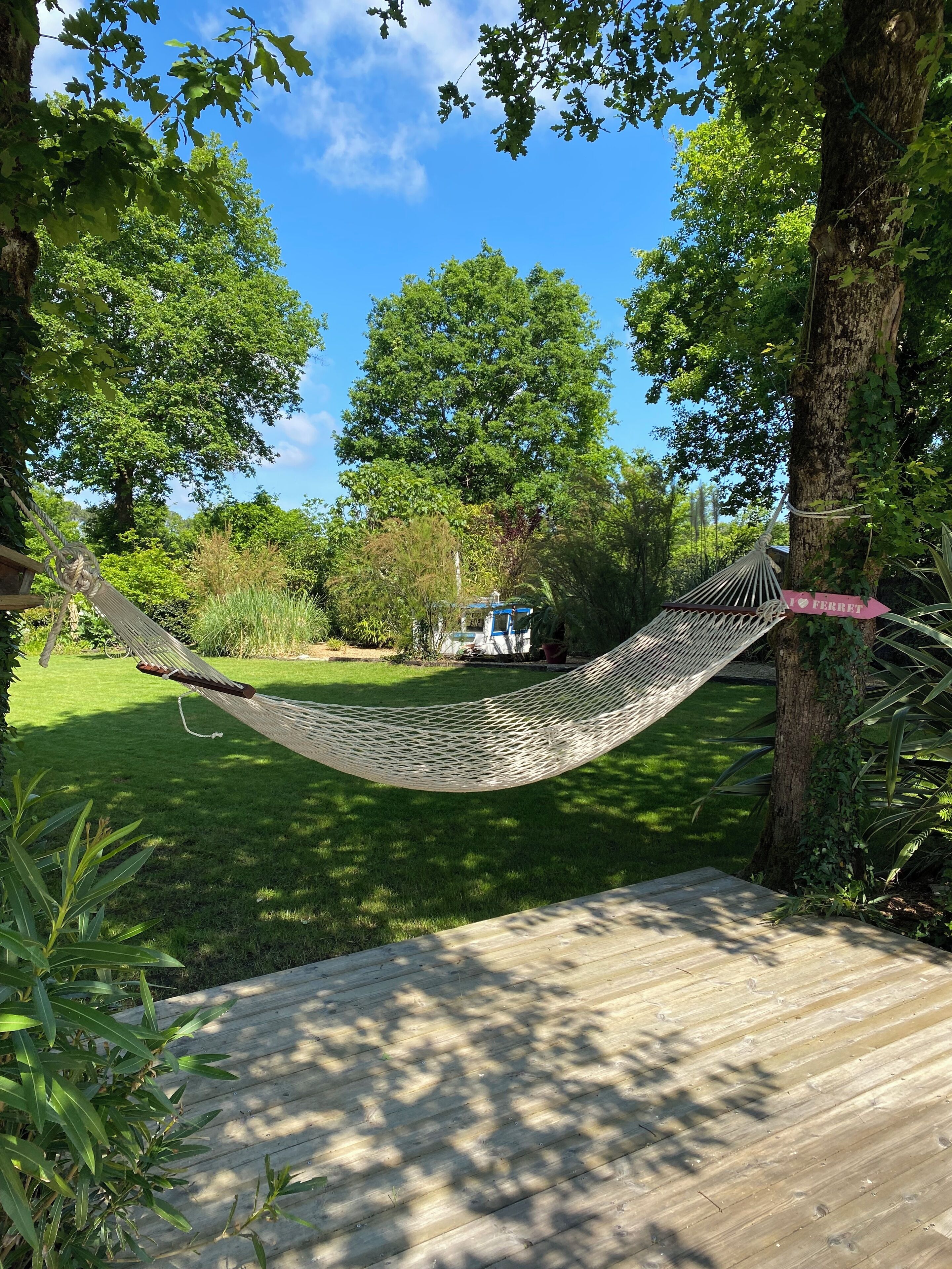 A hammock tied between trees in a lush garden with a wooden deck.