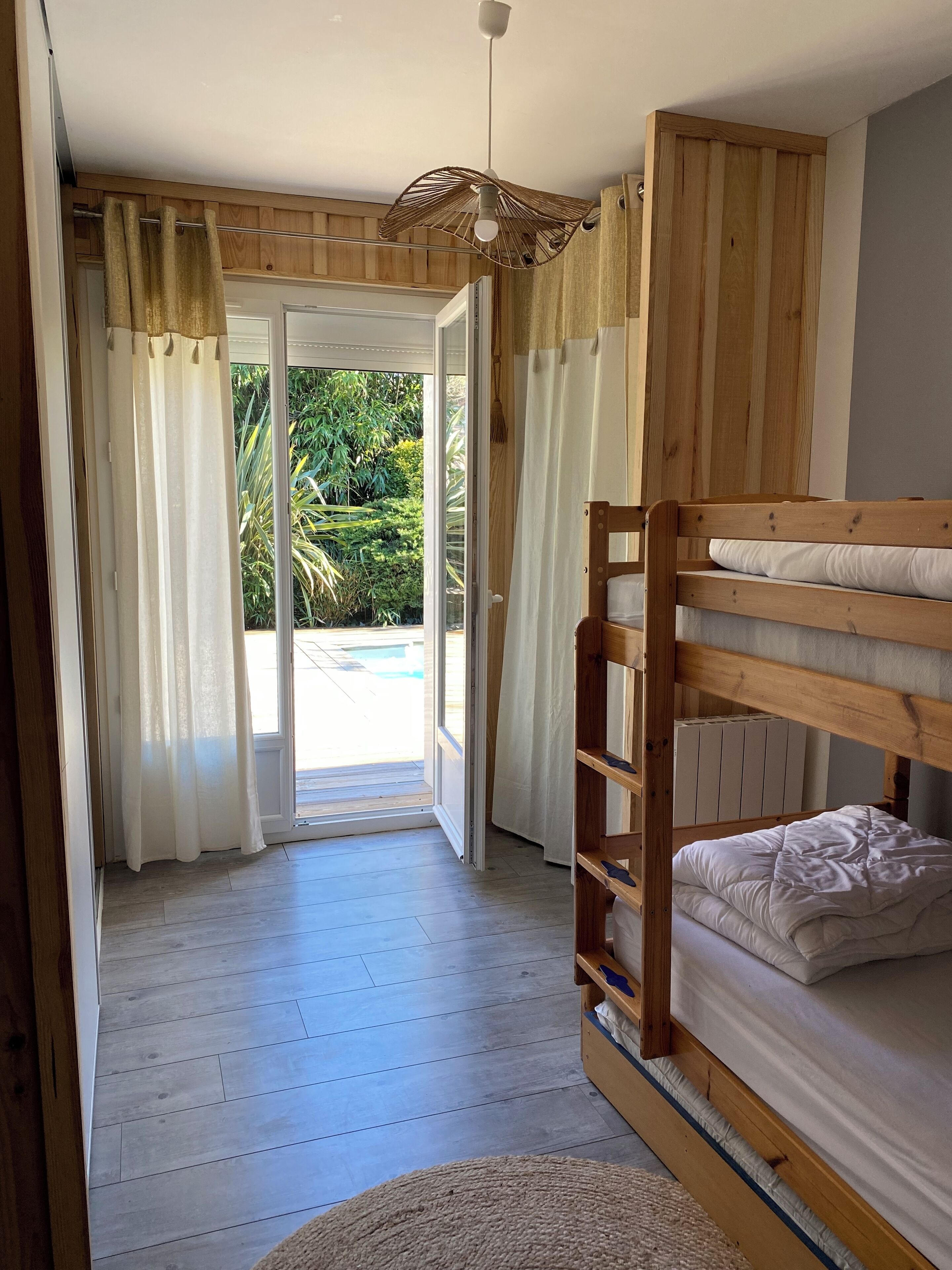 Habitación con literas, piso de madera, ventilador de techo y vistas a una piscina y vegetación.