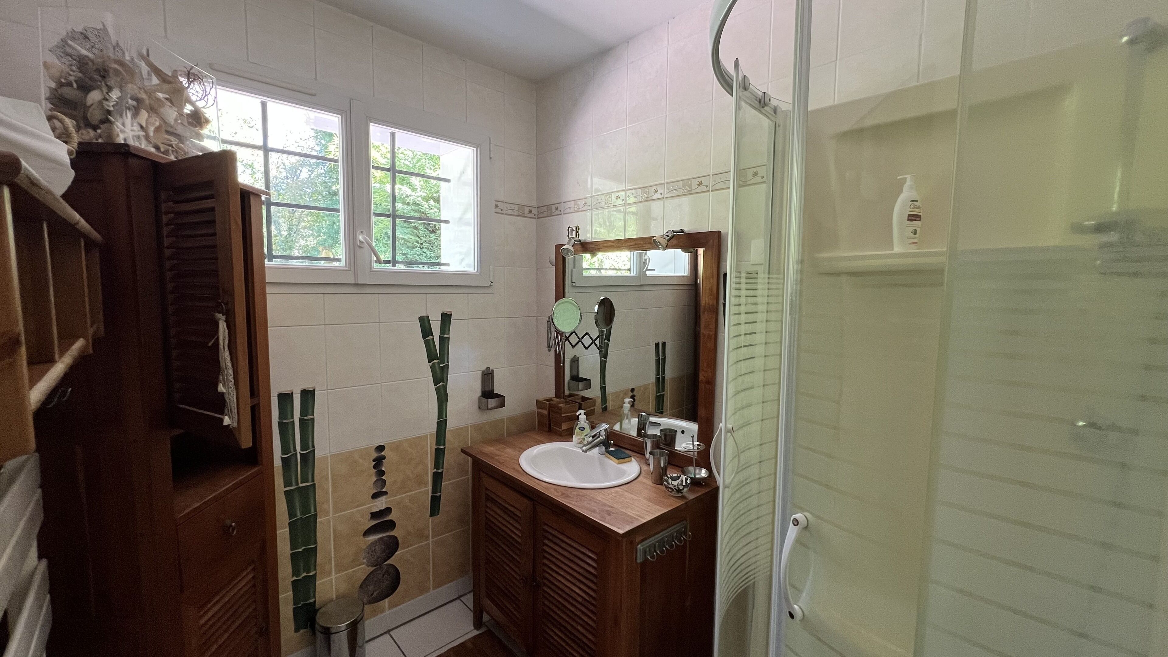 Une salle de bain avec un meuble-lavabo en bois, un grand miroir, une cabine de douche en verre et une fenêtre laissant entrer la lumière naturelle.