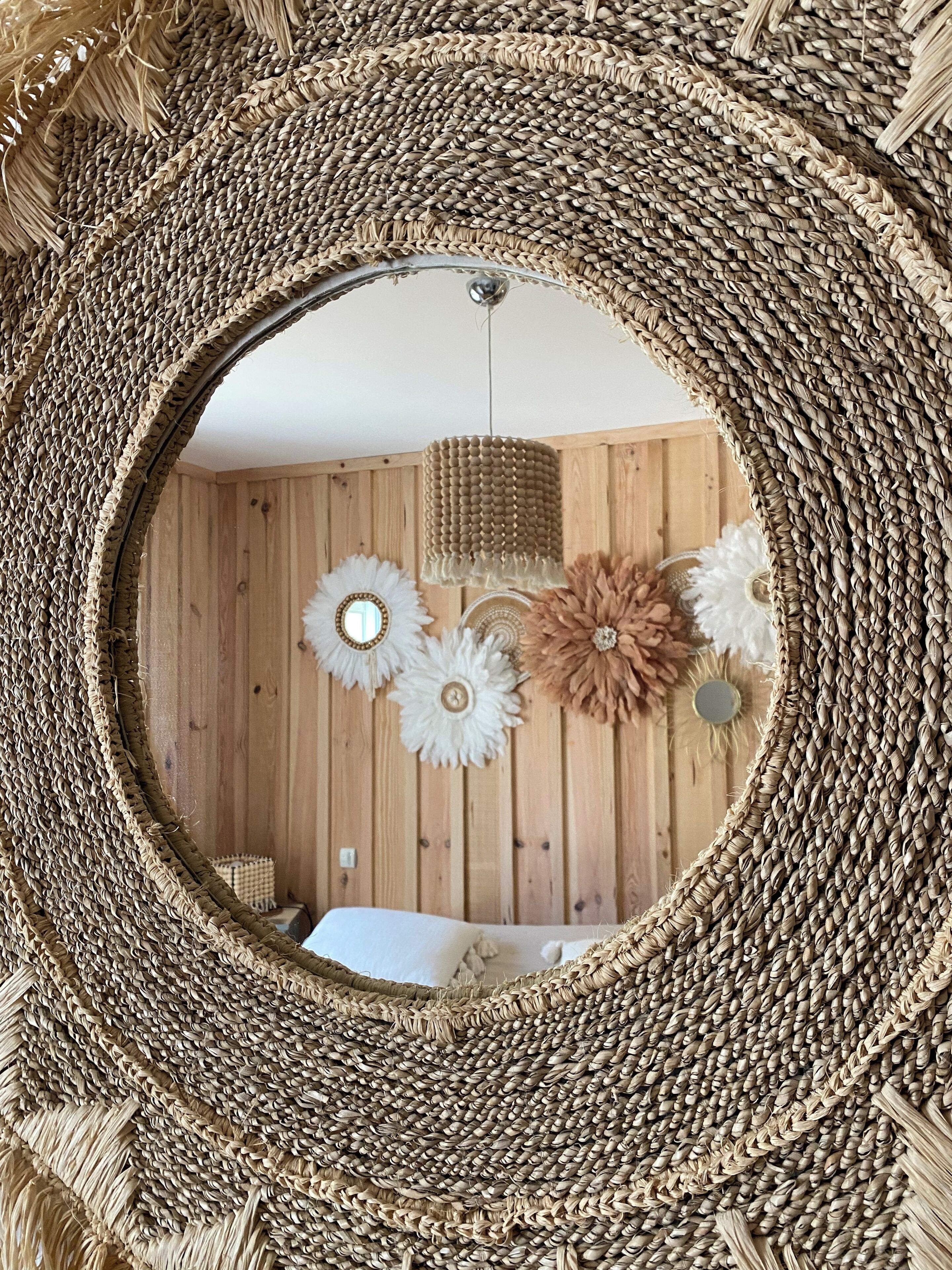 Een ronde spiegel met een geweven lijst, die een kamer weerspiegelt met houten wanden, een bed en decoratieve bloemen.