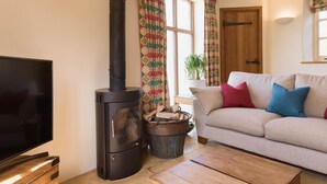 A cozy living room with a black wood stove, a television, a sofa with colorful cushions, and a wooden coffee table. - Cottage 1 - sleeps 4 guests  in 2 bedrooms (New Yatt)