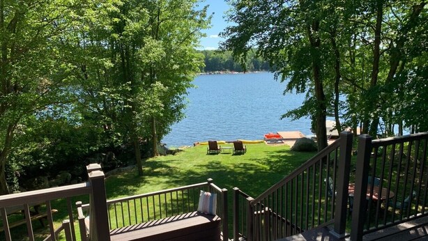 A deck with a railing overlooking a lake, a grassy area with chairs, and a distant shoreline. - SPECTACULAR LAKEFRONT HIDEOUT RESORT-A/C- SWIM+ GOLF-KAYAK-ROWBOAT-BEACH (Hamlin)