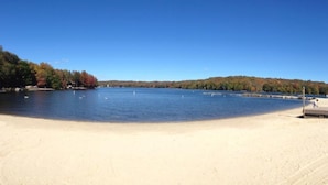 A beach with a sandy shore, a calm lake, and a forested hill in the background. - SPECTACULAR LAKEFRONT HIDEOUT RESORT-A/C- SWIM+ GOLF-KAYAK-ROWBOAT-BEACH (Hamlin)