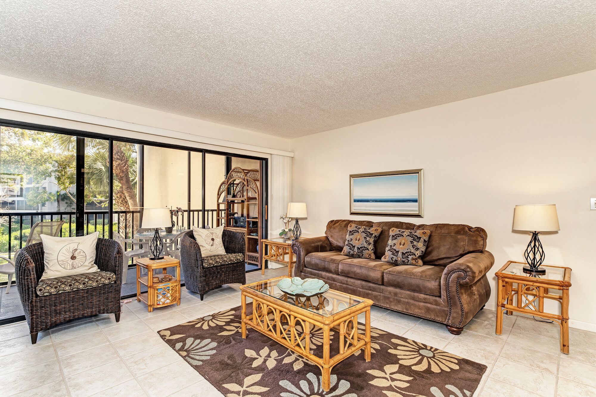 A living room with a brown leather sofa, wicker chairs, a glass coffee table, and a large window offering a view of palm trees.