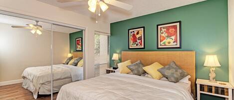 A bedroom with a large bed, a ceiling fan, two bedside tables with lamps, and two framed floral paintings on the wall.