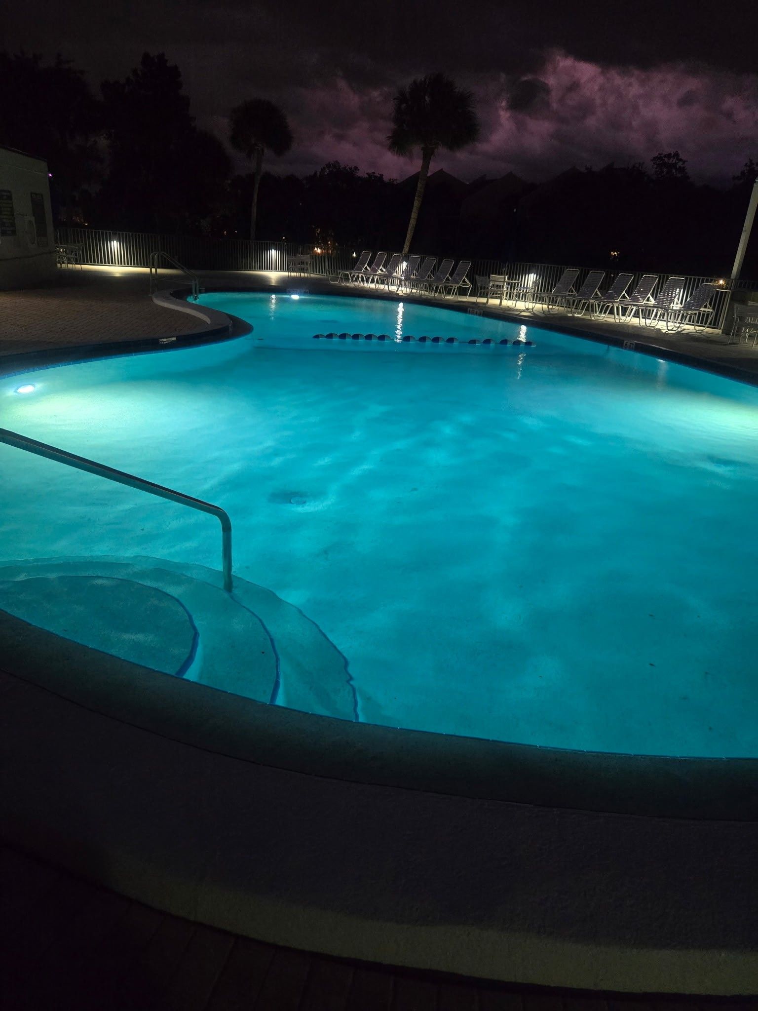 A swimming pool at night with a curved edge, a pool ladder, and lounge chairs along the perimeter.