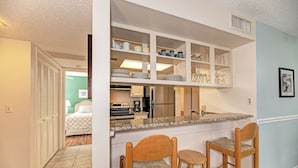 A compact kitchen with a built-in island, a refrigerator, and a coffee maker. There are two wooden bar stools and a bed visible in the background. - Beautiful Siesta Key Condo (Siesta Key)
