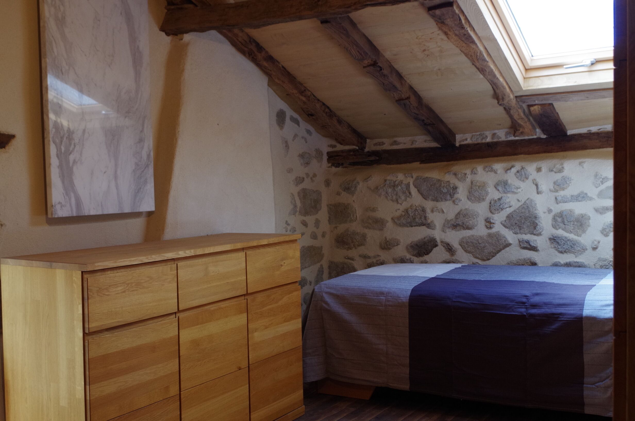A bedroom with a wooden dresser, a bed with a blue blanket, stone walls, and a skylight.