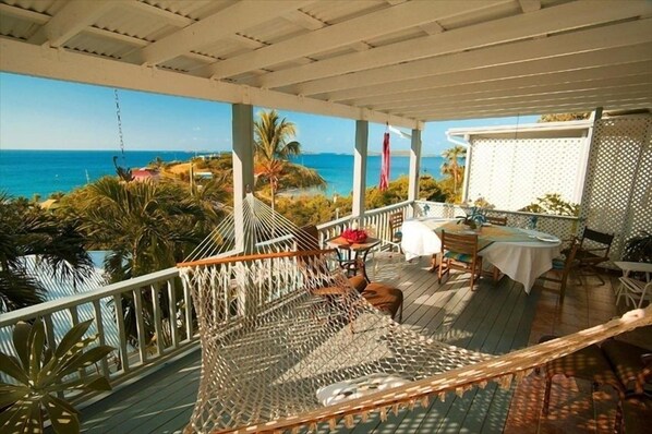 A covered outdoor area with a hammock, tables, and chairs, overlooking a beach and ocean.