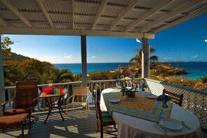 A covered outdoor dining area with a table set for two, overlooking a coastal landscape with clear skies.
