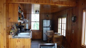 Interior de una cabaña de madera con zona de cocina que incluye un lavamanos, estufa, refrigerador y mesa de comedor con sillas.