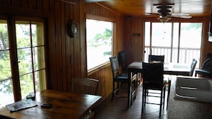 Interior de una cabaña de madera con una mesa de comedor, sillas, una zona de cocina y ventanas amplias.