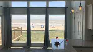 A room with a large window overlooking a beach, a table with a rose and wine glasses, and a hanging light fixture. - Beach Front on Galveston's West Beach (Galveston)