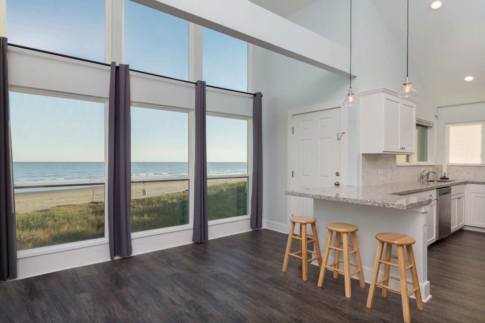 Ett modernt kök med stora fönster som vetter mot en strand, trästolar och en bänkskiva i granit.
