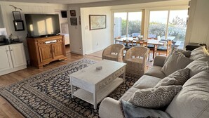 Uma sala de estar ampla com um sofá grande, uma mesa de centro e um espaço de jantar com cadeiras e uma mesa. Há uma área de cozinha com armários e uma televisão.