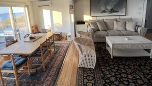 Uma sala de estar espaçosa com uma janela grande, uma área de jantar com móveis de madeira, um conjunto de sofás e um tapete estampado.