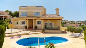 Una casa de dos pisos con piscina, un patio con sillones de descanso y vistas del entorno.