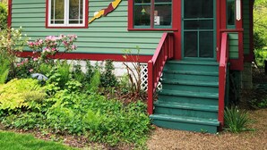 A green house with a red door, a small porch, and a garden with various plants. - Lakeside Grove Cottage -  Perfect Lakeside Village/Beach Location (Lakeside)