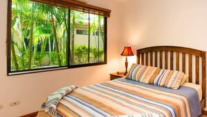 Ein Schlafzimmer mit Bett, gestreifter Bettwäsche, einem Nachttisch mit Lampe und einem Fenster mit Blick auf Grünflächen.