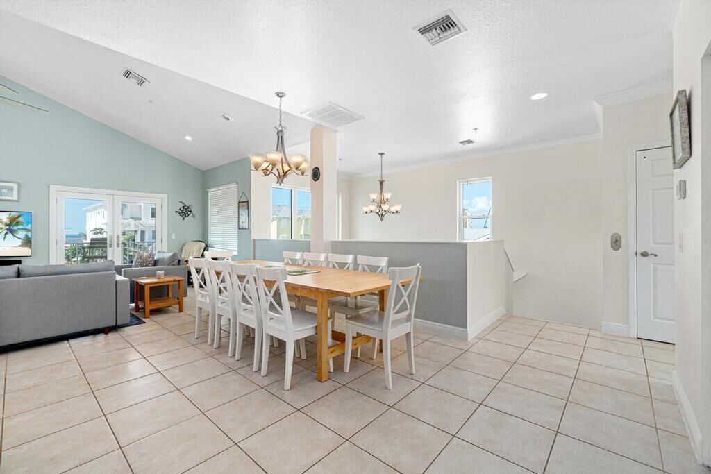 A modern kitchen with a dining area, featuring a wooden table and white chairs, a grey sofa, and a view of the outside.