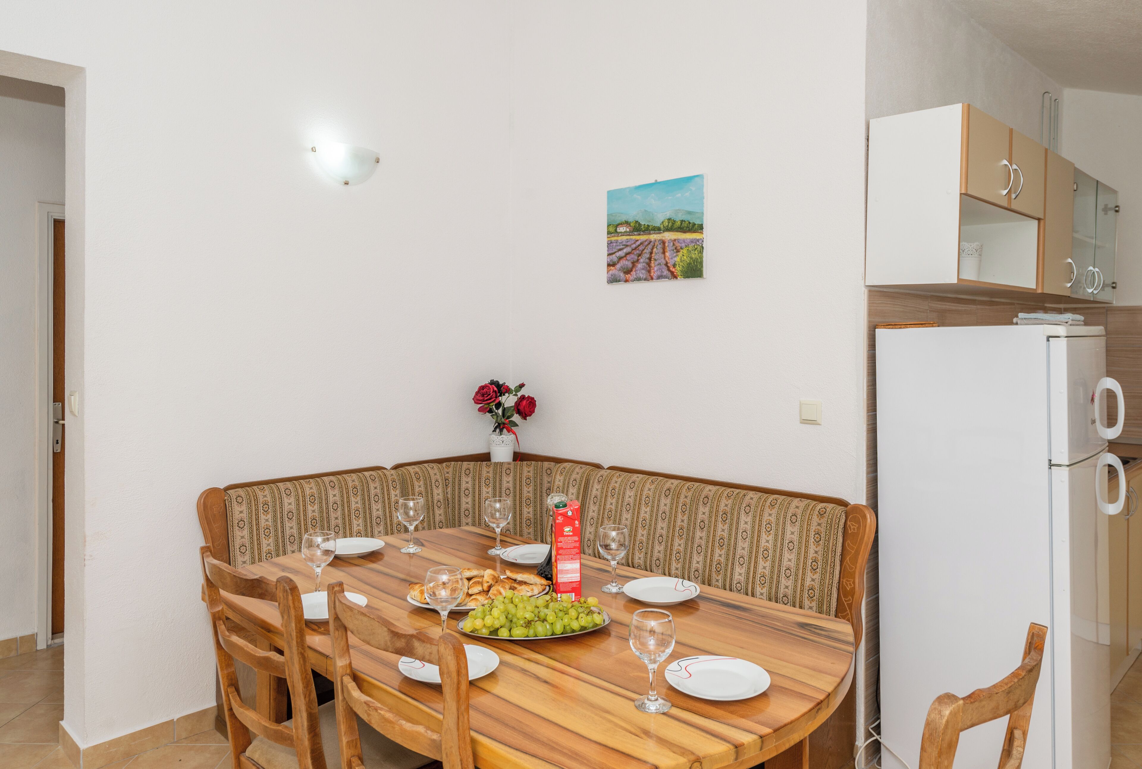 Een eethoek met een ronde houten tafel, stoelen en een bank. Er staan borden, glazen en een doos wijnglazen. Op de tafel staat een vaas met rode bloemen.