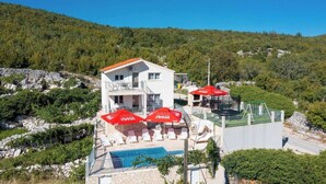 A two-story building with a pool area, red umbrellas, and a mountainous backdrop. - Holiday house Marijana (Zagvozd)