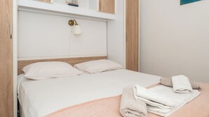 A neatly made bed with a pink blanket and rolled towels, a white wall with a small shelf and a wall-mounted lamp, and wooden paneling on the walls. - Holiday house Marijana (Zagvozd)