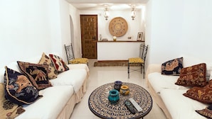 Salón con dos sofás blancos, cojines estampados, una mesa redonda de centro con objetos decorativos y una lámpara fijada a la pared.