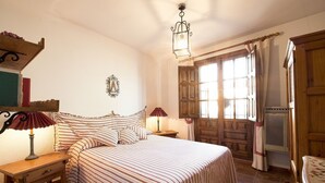 Un dormitorio con cama, dos mesitas de noche, lámpara, ventana con cortinas, puerta de madera y aplique de pared.