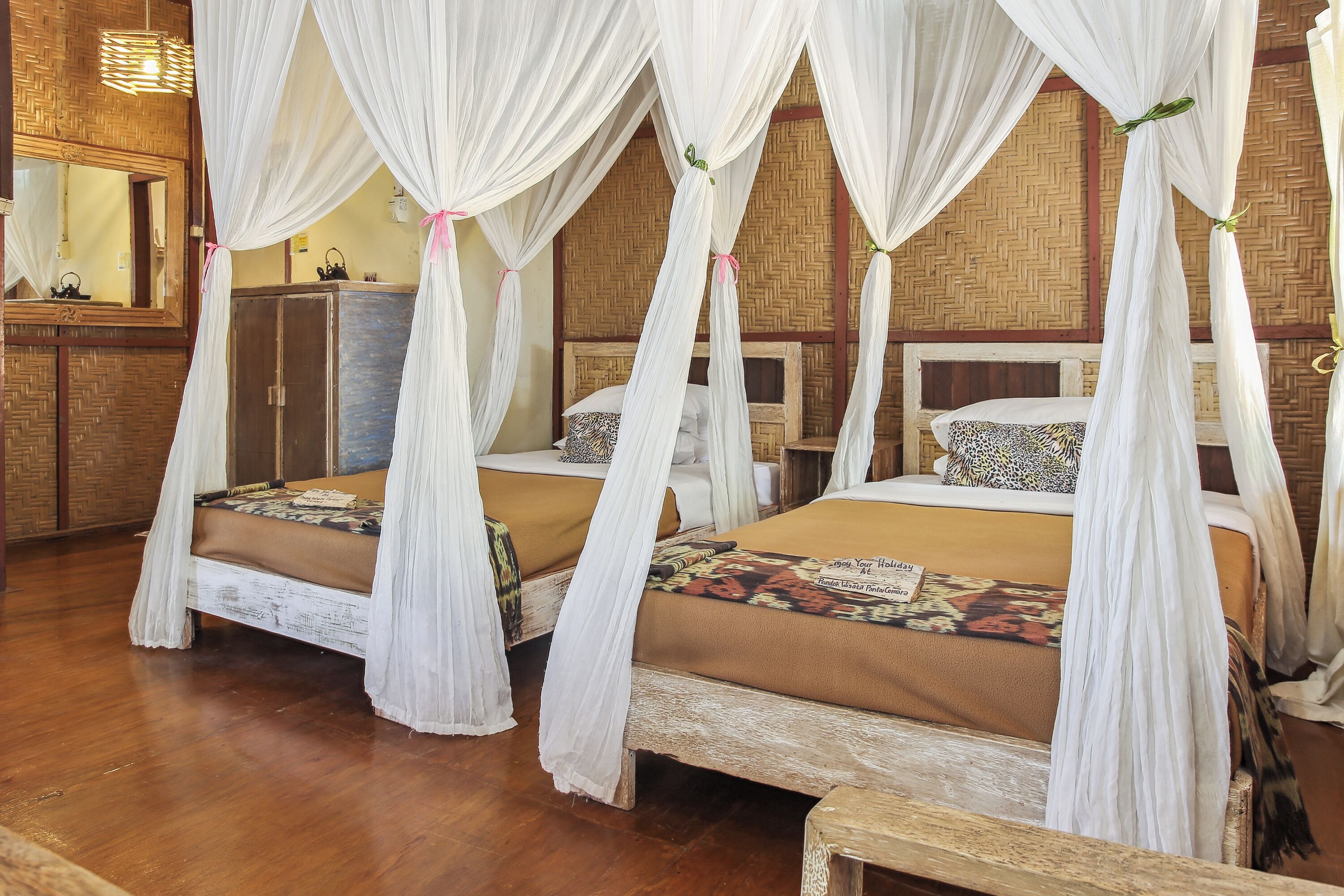 A room with two beds, each with a canopy bedspread and a headboard. There is a wooden bench in the foreground, and a mirror on the wall.