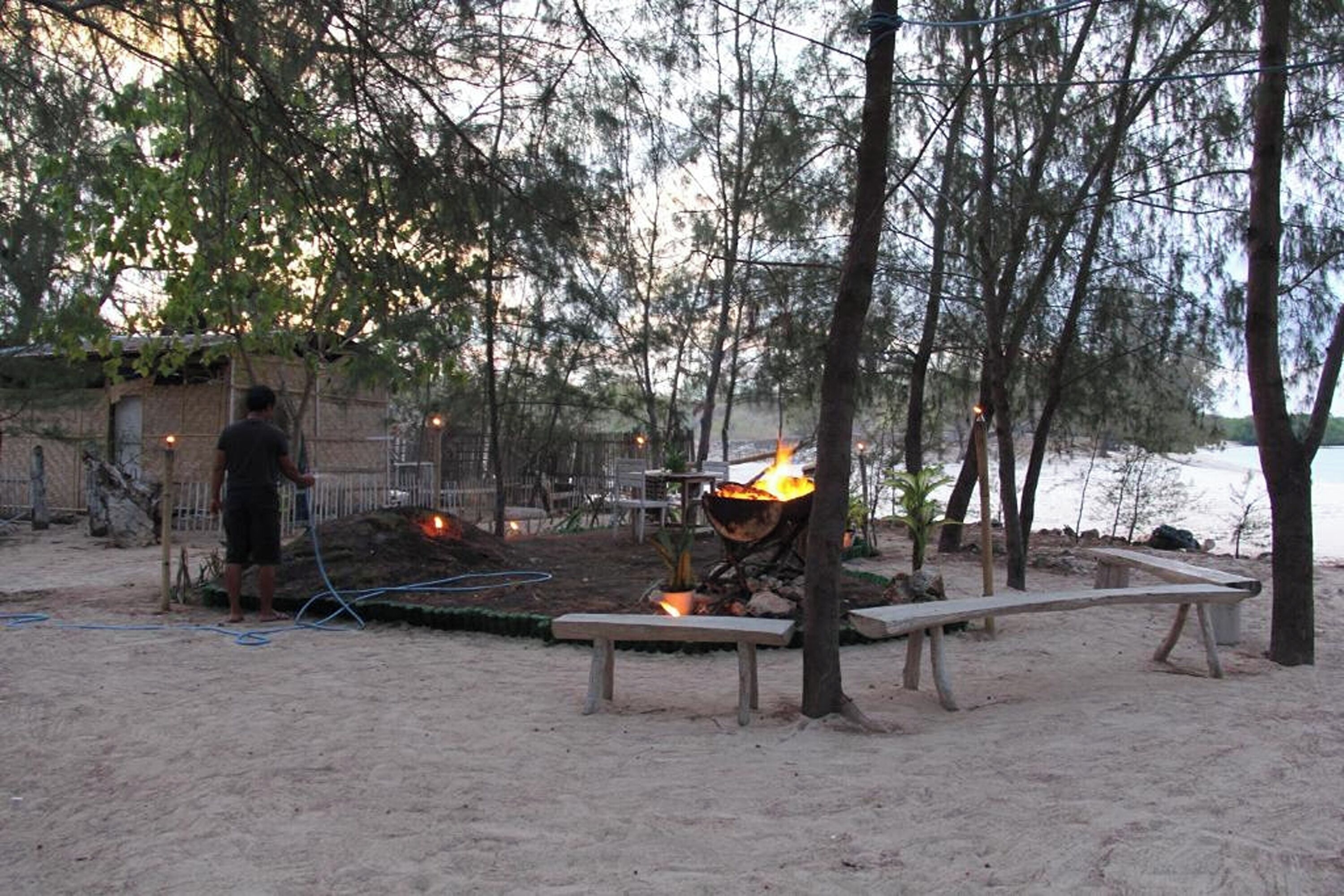 A beachside outdoor area with a fire pit, wooden benches, and a person standing nearby.