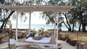Uma cama de madeira com roupa de cama branca e duas almofadas estampadas, colocada numa praia arenosa com uma pérgula de madeira por cima.