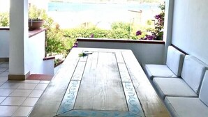 Une terrasse couverte, dotée d’une table en bois et de sièges blancs rembourrés, offre une vue panoramique sur un plan d’eau et une végétation luxuriante.