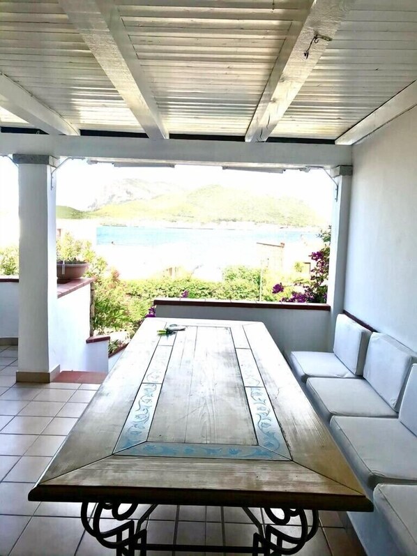 A covered patio with a wooden table and white cushioned seating, overlooking a scenic view of a body of water and greenery.