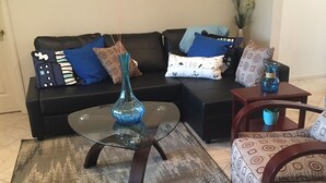 A living room with a black sofa, patterned armchairs, a glass coffee table, and a painting of boats on the wall.