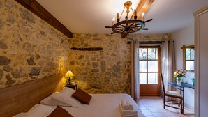 Un dormitorio con pared de piedra, cabecera de madera, cama con ropa blanca, una silla de madera y un escritorio con un florero.