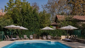 Piscina con sillas de descanso y sombrillas, rodeada de vegetación y un edificio de piedra.